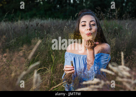 A pretty young woman in her twenties blows golden glitter from her hand during an outdoor portrait shoot one evening near Liverpool. Stock Photo