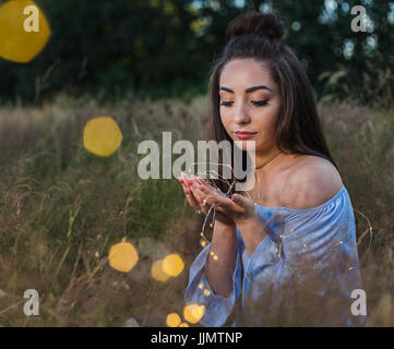 A beautiful young woman is wrapped in fairy lights whilst posing for a portrait in an area of wild grass near Liverpool. Stock Photo