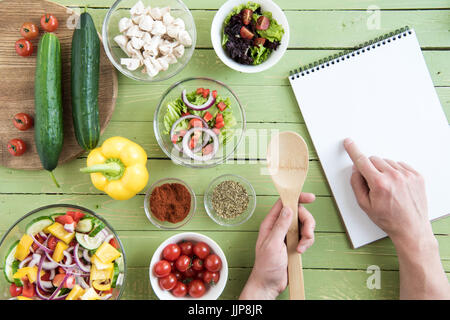 Close-up partial view of person holding wooden spoon and pointing at recipe in cookbook while cooking Stock Photo