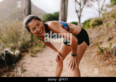 Fit young asian woman standing on mountain trail with her hands on knees and smiling. Female runner in sportswear taking a break after running workout