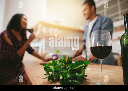 Glass of red wine on kitchen counter with couple standing in background. Man and woman talking in the kitchen with glass of wine in front. Stock Photo