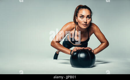 Horizontal shot of young fit woman doing push up on medicine ball. Fitness female exercising with a medicine ball on grey background. Stock Photo