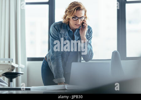Businesswoman talking on cellphone and looking at laptop. Female executive working in office. Stock Photo
