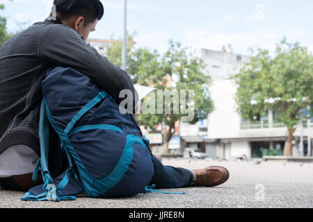 young traveler reading map. handsome asian man wearing black jacket and blue jeans sitting in park with backpack Stock Photo