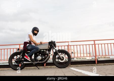 Handsome happy rider guy in red sneakers, white t-shirt and black moto helmet sit on his classic style biker cafe racer motorcycle. Bike custom made i Stock Photo