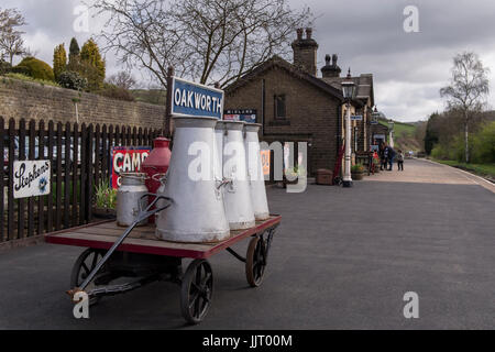 View of old adverts & milk churns on cart, on platform at quiet, quaint, historic Oakworth Station - Keighley and Worth Valley Railway, England, UK. Stock Photo