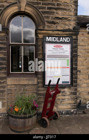 View of red porters' trolley by train times poster on platform at quaint, historic Oakworth Station - Keighley & Worth Valley Railway, England, UK. Stock Photo