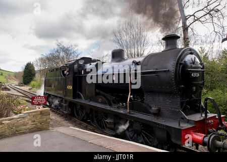 Engine driver in steam locomotive puffing smoke (BR Midland Railway 4F 0-6-0 43924) pulls into station - Keighley & Worth Valley Railway, England, UK. Stock Photo