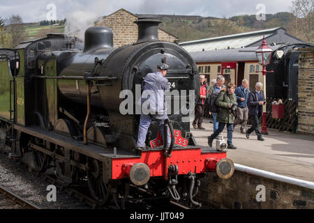 People on platform walk past train driver climbing on front of steam locomotive BR 4F 43924 - Keighley and Worth Valley Railway station, England, UK. Stock Photo