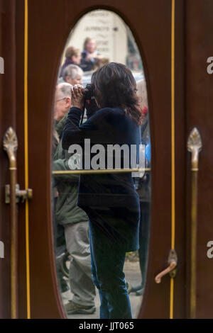 Lady with camera taking train photo, seen through glass door of old-fashioned Pullman carriage - Keighley & Worth Valley Railway station, England, UK. Stock Photo