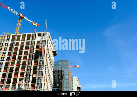 Low angle view of two concrete buildings under construction next to a glass building with two tower cranes against blue sky. Stock Photo