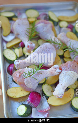 Baking sheet with chicken legs, zucchini, rosemary, onions, and potatoes before going in the oven Stock Photo