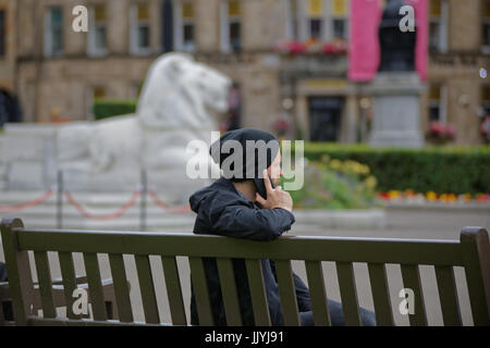 young man male student teenager  on telephone mirrored by lion statue in the background from behind  or side on sitting om bench Glasgow George Square Stock Photo