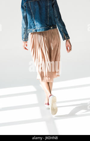 Rear view, low angle shot of woman in beige skirt and denim jacket walking in white studio Stock Photo