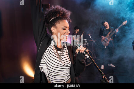 Female singer with microphone and rock and roll band performing hard rock music on stage Stock Photo