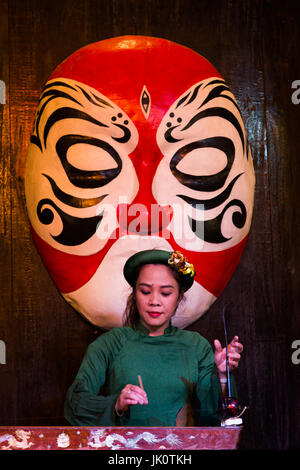 Female folk musician playing traditional instrument in front of painted mask - Hoi An - 14 March 2017 Stock Photo