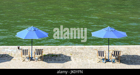 Two blue parasols with blue and white striped deck chairs in the sun on the wharf of the river Seine with green water. Stock Photo