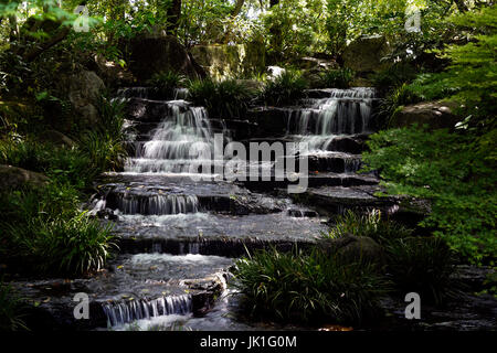 Small waterfall streaming down black rocks in between green vegetation. Stock Photo