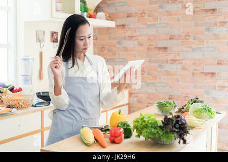 Cheerful Asian young woman is cooking in the kitchen with joy. She is standing and holding digital tablet of recipe. Asian woman is touching a wood