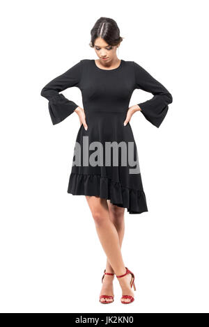 Sad pretty woman in black dress with hands on waists looking down. Full body length portrait isolated on white background. Stock Photo