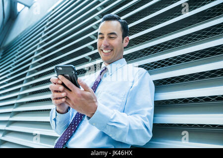 Smiling Mixed Race businessman leaning on wall texting on cell phone Stock Photo