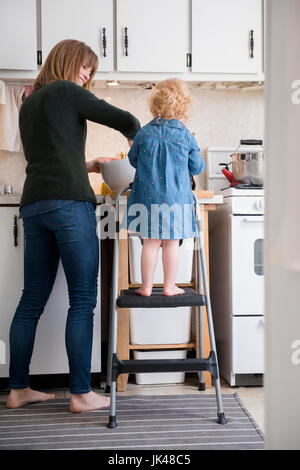 Caucasian girl standing on ladder cooking with mother in kitchen Stock Photo