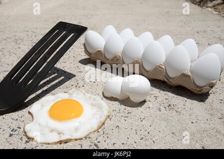 Fried egg with broken shells and carton of white eggs on hot sidewalk on summer day. Stock Photo