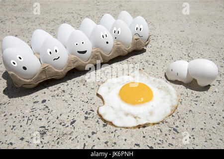 Fried egg with broken shells and carton of white eggs with illustrated faces on hot sidewalk on summer day. Stock Photo