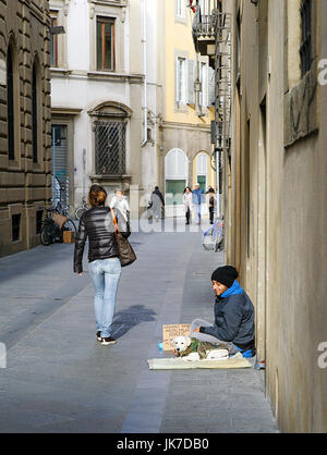 Florence, Italy: November 14, 2016: A young male beggar sits on the ground in an alleyway, with his dog beside him. A young woman walks past and other Stock Photo