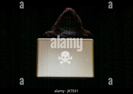 An anonymous figure with a hooded sweatshirt types on a laptop surrounded by a green code matrix, with a sinister skull as logo on laptop, hacking Stock Photo