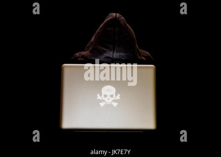 A hacker, shrouded in anonymity, works on a laptop in the dark, with a sinister skull as logo on laptop, hacking and cybersecurity concept Stock Photo