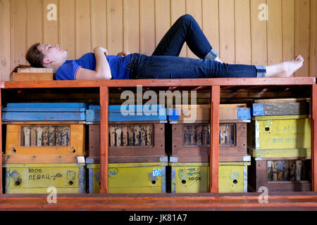 A young Siberian woman laying over a stack of wooden Langstroth hive with active honey bees in a Bee Therapy Hut in the mountainous region of SEZ TRT 'Biruzovaya Katun' located in Altai Krai a region in Western Siberia, Russia. Beehive air is highly-ionized and clean, which has been shown to benefit the lungs and respiratory system as well as have a calming effect on the mind.
