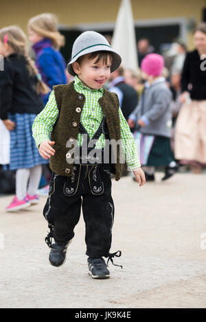 Child in traditional Bavarian dress with leather trousers, cardigan and fur hat sticking his tongue out walking on the dance floor around a maypole Stock Photo