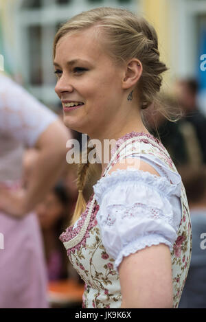 Young Bavarian woman in traditional dirndl dress with arms akimbo looking aside while performing traditional folk dance Bandltanz around the maypole Stock Photo