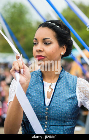 Young woman in traditional dirndl dress holding white ribbon while performing traditional folk dance Bandltanz around the maypole Stock Photo