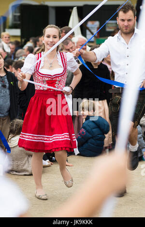 Young Bavarian woman in traditional dirndl dress holding blue ribbon while performing traditional folk dance Bandltanz around the maypole Stock Photo