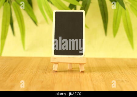 Small empty slate in height to write a message that is posed on an easel all on wooden floor and green foliage background Stock Photo