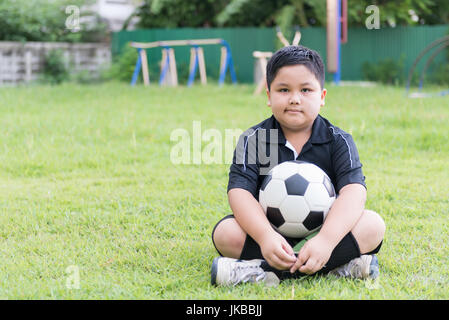sitting obese fat boy  soccer player with football on grenn grass background, healthy concept Stock Photo