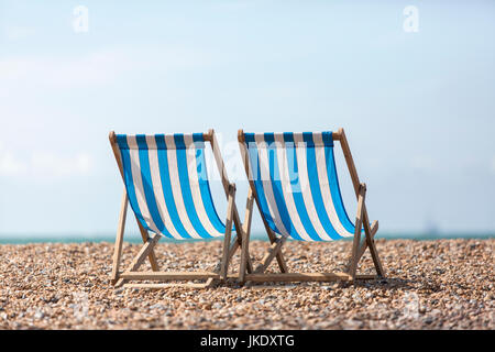Two empty blue and white striped deck chairs facing out to sea, on a pebble beach.
