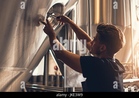 Brewer closing the hatch of brewery tank. Young male employee working in beer manufacturing factory. Stock Photo