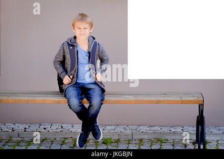 Boy with backpack sitting on the bench near white mock up poster on school wall Stock Photo