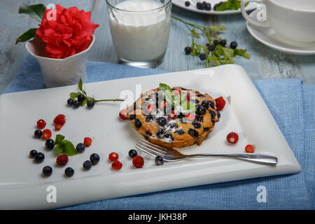 Berry muffin with blueberries and strawberries on a white plate and a wooden table served with milk and pink rose in a vase Stock Photo