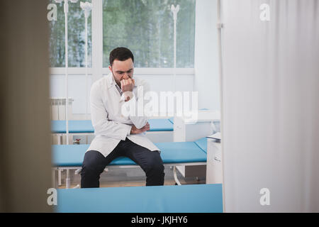 Handsome young doctor in white coat is sitting sadly in hospital ward Stock Photo