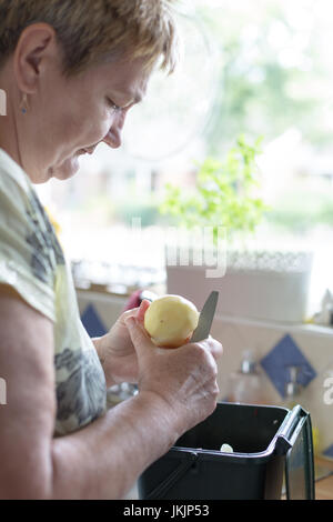 Senior woman cuts peeled potatoe, preparing food. Vertical orientation with selective focus on potatoe and knife. Natural day light Stock Photo