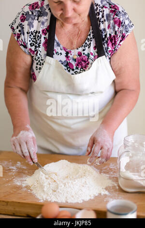Senior woman makes dough using flour and pouring water. Top point of view, selective focus on working hand with knife. Vertical crop Stock Photo