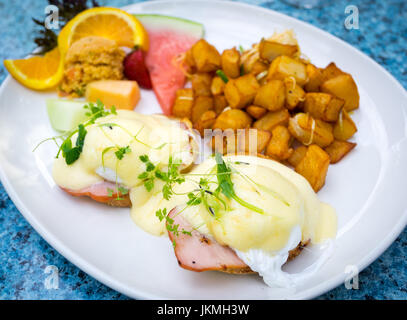 Classic Eggs Benedict (soft poached eggs, Canadian back bacon, and Hollandaise sauce on an English muffin), fried potatoes, and fruit for breakfast. Stock Photo