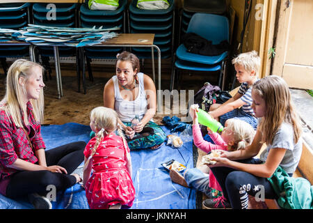 Caucasian adults and children sitting on floor in circle chatting to each other at indoor creative arts and crafts workshop. Stock Photo