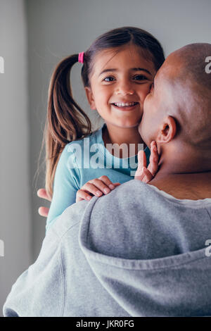 Little girl laughing and looking at the camera. Her father has his back to the camera and is holding the girl while kissing her face. Stock Photo