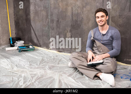 Handsome young laborer sitting on floor with digital tablet and smiling at camera Stock Photo