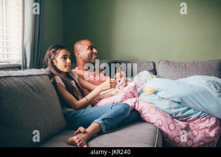 Father curled up on the sofa with his two daughters. They are watching a movie with popcorn. Stock Photo